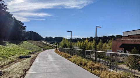 A photo of a long paved walking and jogging path with bridges, beneath blue skies and beside a bank of trees. 