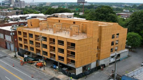 An aerial photo of a plywood building near many trees under gray skies. 