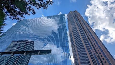 A photo of a glass flat-fronted new tower in Atlanta under blue skies next to a huge brown tower. 