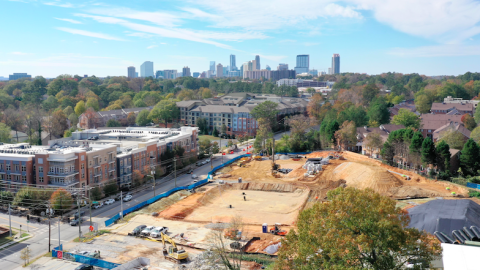 A drone image of a large city with a new construction project of beige dirt at the bottom of the photo amongst many multicolored trees.