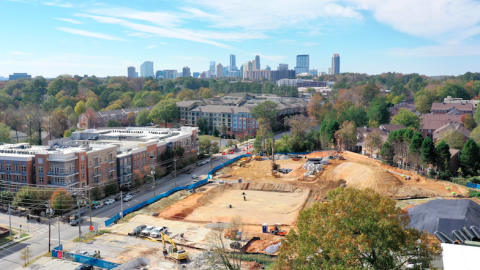 A drone image of a large city with a new construction project of beige dirt at the bottom of the photo amongst many multicolored trees. 