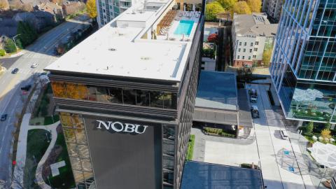 A photo of a new black-clad Nobu hotel near many new buildings under blue skies. 