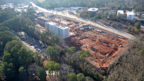 An overview photo of a huge construction site of red dirt and new white buildings beside a wide highway in Atlanta, near trees.