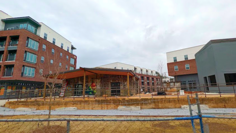 Under gray skies, a new collection of brick buildings with a small old building in front near a dirt walking path. 