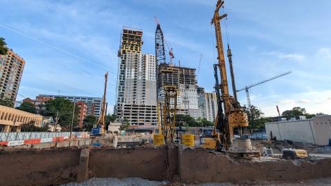 A construction site under blue skies showing a large how and much heavy equipment near new tall buildings. 