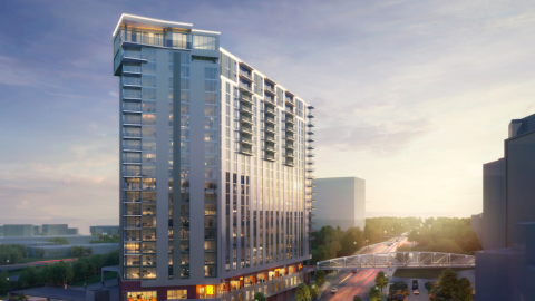 A glassy apartment tower with shops at the base shown at daybreak near a stadium.