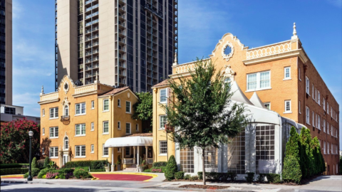 A grand spanish style yellowish orang hotel under blue skies with a tall building behind.