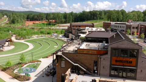 A cluster of man brick buildings beside a forest under blue skies with a large green amphitheater and a fountain included in new development.