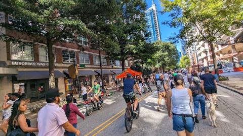 A photo of people on bikes and walking beside many tall buildings under blue skies in an open street.