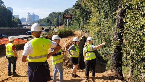 A dirt path under construction near many trees and woods and a wide highway. 