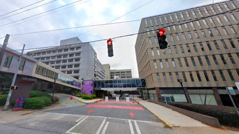 An image of a hospital under blue-gray skies with a large road in front near stoplights. 