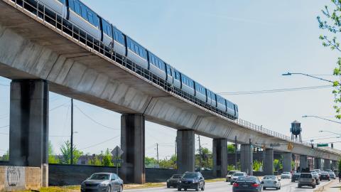 An image of a long metal train on elevated tracks over a wide road under blue skies.