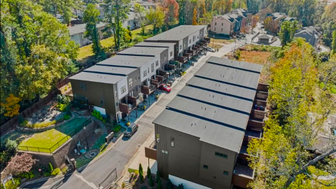 A photo of a large row of black and white and wood-clad townhomes with white modern-style interiors on a lane behind a gate with many trees.