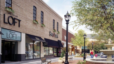 A photo showing a large fountain next to a row of shopfronts and wide sidewalks and trees.