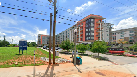 An image showing a large apartment building under blue skies next to a wide road and green grassy field.