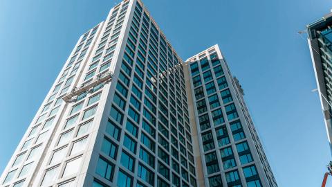 Photos showing a large glass and stucco new tower under blue skies with small white apartments inside.