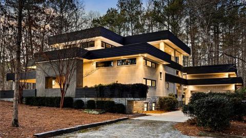 A photo of a large stone and concrete modern home in a wooded setting with black trim and modern white interiors with many windows.