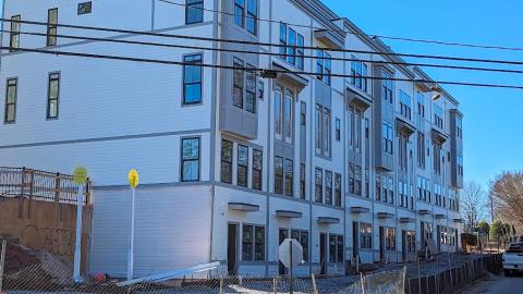 A photo of a large new townhome and condo project in white and gray under blue skies near a wide street.