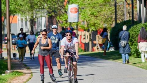 An image of many people riding and walking on a wide sidewalk area beside bushes. 