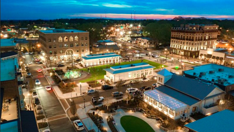 A photo of a large suburban city center with a greenspace in the middle and many buildings new and old around it.