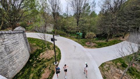 A photo of a wide new concrete trail built in woods with lampposts and people jogging and biking on the path.