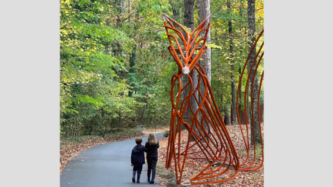 A photo of a large metal new sculpture in a wooden setting near a wide pathway with people walking on it in Atlanta.