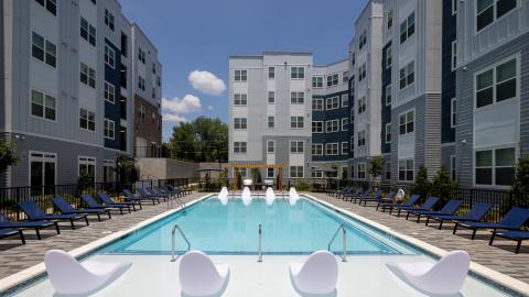 An image of a new grey, blue and white apartment complex under blue skies with modern interiors and a pool with a sun ledge in the middle.