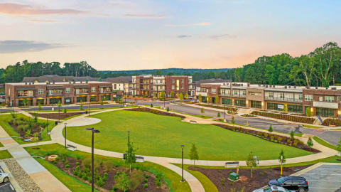 A photo of a large apartment community near a forest with a green in the middle and shops and apartments with white modern interiors.