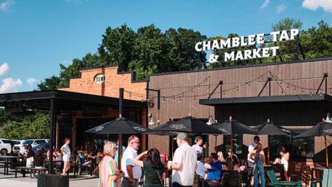 A photo of many people with drinks hanging out outside a new brick building with a sign atop it under blue skies. 