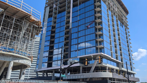 A photo of a large glassy building that's topped out with a crane above, with another rounder building under construction next door. 