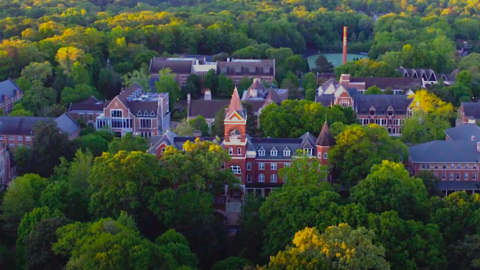 A photo of a large gothic brick building on a college campus with many trees surrounding it, with modernized new interiors.