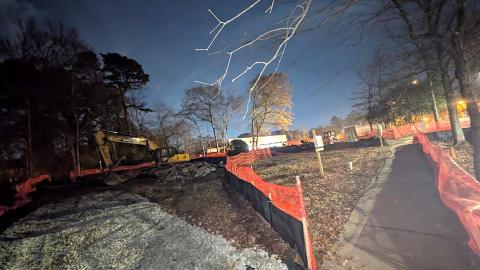 A large construction site shown under bare trees under night skies near a large shopping center.