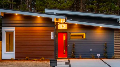 A photo of a new house with midcentury modern style and a red door and blue siding near many trees.