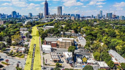 An overview of a retail lot where a low building stands today with many trees and homes around and tall buildings in the distance in Atlanta.