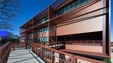 A modern brown and tan new mid-rise structure on a college campus with many leaves and trees, under blue skies.