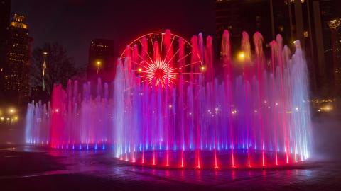 A photo of a large fountain shaped like Olympic Rings with many lights near skyrises in a city.