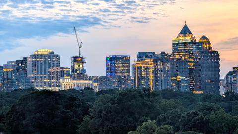 An evening photo of the Atlanta skyline with a pink, orange, and blue sunset and many buildings over trees.