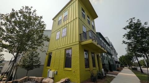 A photo of a row of stacked buildings painted green gray and white under hazy blue skies near wide streets and trees.