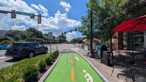 A photo of a long protected bike lane near a wide street through a city and college campus under blue skies with fluffy clouds.