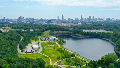 A drone photo of a huge park with a large lake and sculptures with many green trees and a big city beyond.
