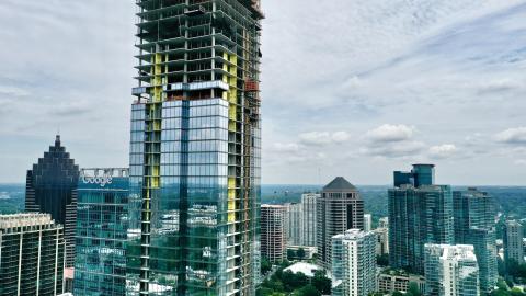 A drone photo of a very tall glassy new building with a large yellow crane at top in Midtown Atlanta under blue gray skies with many other high-rises standing around.