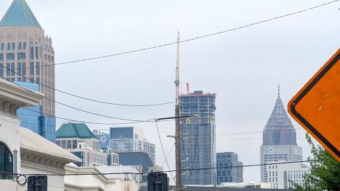 A photo of a large glass and concrete high-rise climbing in Atlanta near many buildings under gray skies, along a wide busy street. 