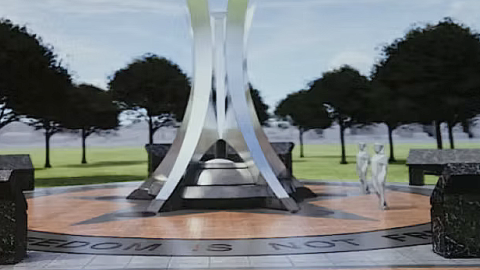 An image of a site in a park with a large steel memorial and wall with many names, with a brick walkway and trees dotted around.