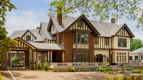A photo of a large Tudor mansion in Atlanta under blue skies with large trees and a white new building beside it.