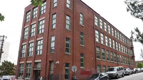 A photo of a red brick four-story building on a corner of two streets in Atlanta under gray skies near trees. 