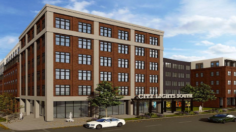 An image of a site for a large brick affordable housing structure under blue skies on a corner where smaller buildings stand just east of downtown Atlanta.