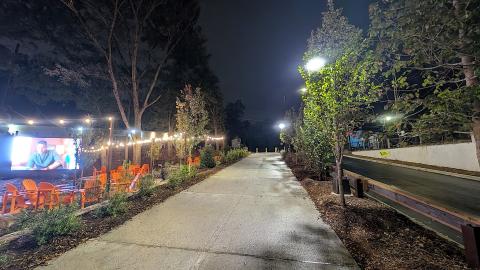 A photo of a wide new biking and pedestrian trail next to a leafy brewery with many trees and lights and a giant TV in Atlanta at night.