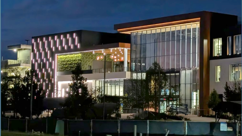 An image of a large new modern complex with much glass and construction fencing under cloudy skies at night.