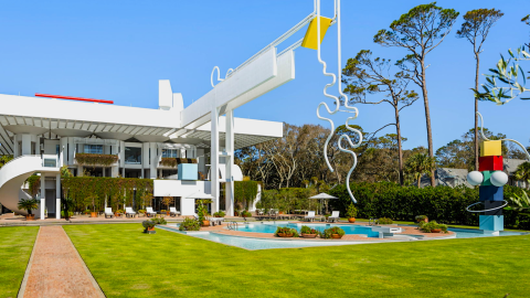A photo of a huge white modernist beach house with big art displays near many trees with airy modern interiors and views of the ocean.
