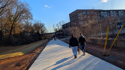 A photo of a new concrete stretch of the Atlanta Beltline that's open under blue skies with many people on it and bridges and trees. 
